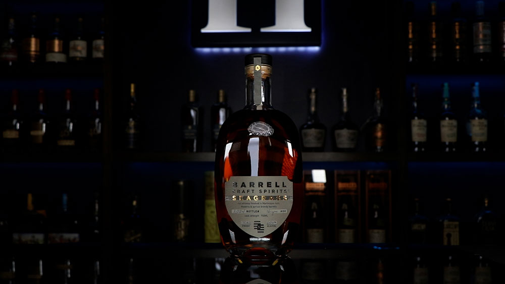 A bottle of Barrell Seagrass 16 Year sitting in front of a Homebar.io light up sign with shelves filled with bottles.
