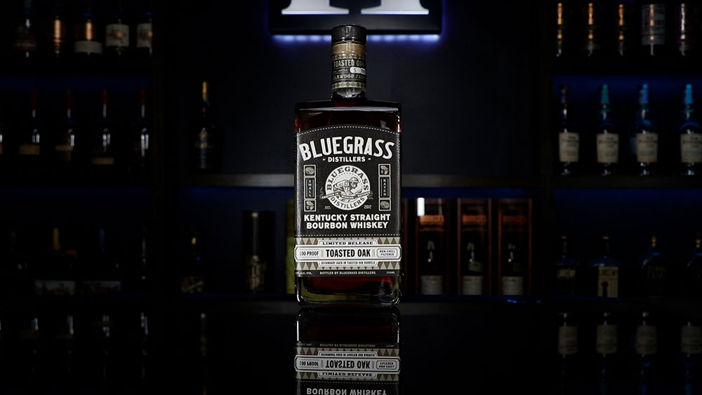 Bluegrass Distillers Toasted Oak sitting on a table in front of shelves filled with bottles.