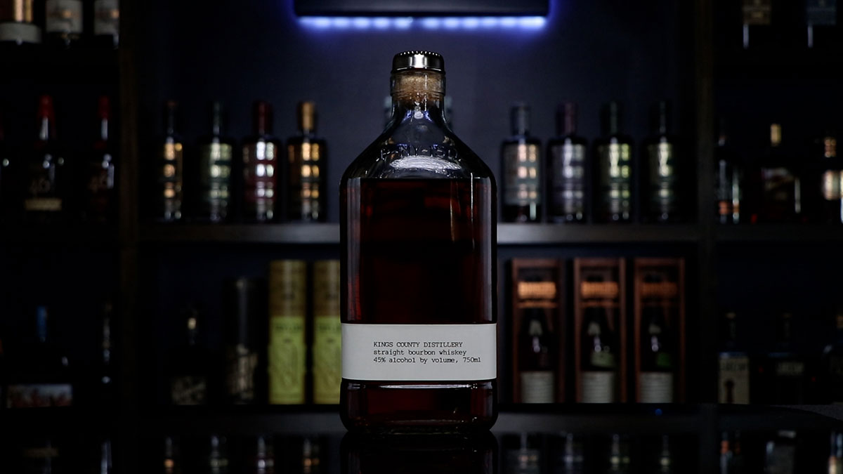 A bottle of Kings County Distillery Straight Bourbon Whiskey sitting in front of a Homebar.io light up sign with shelves filled with bottles.