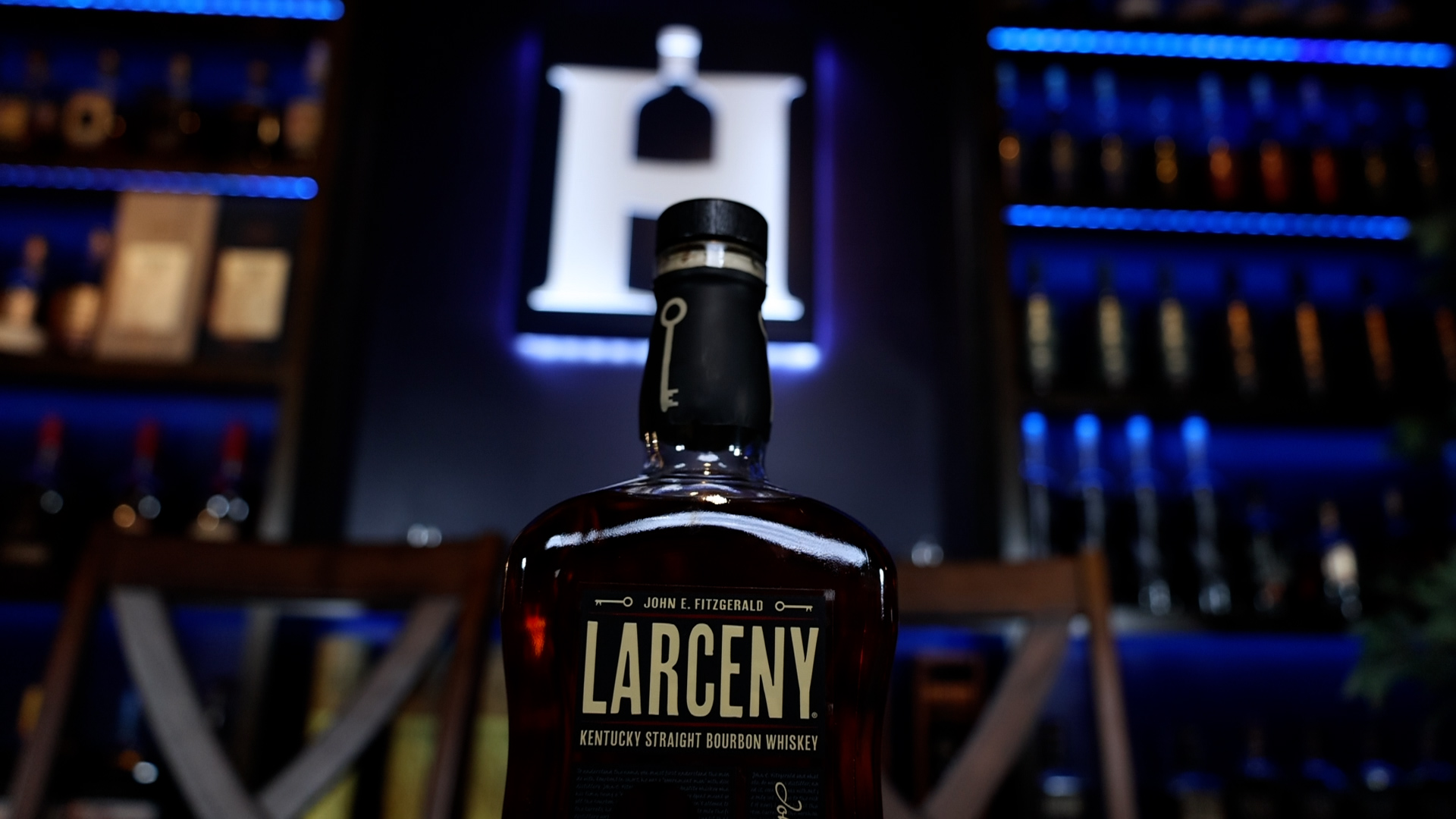 A bottle of Larceny Barrel Proof whiskey sits on a table in front of a lit-up Homebar.io logo sign.