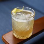 A rocks glass sitting on a wooden armchair containing a cocktail called a 'Gold Rush'.