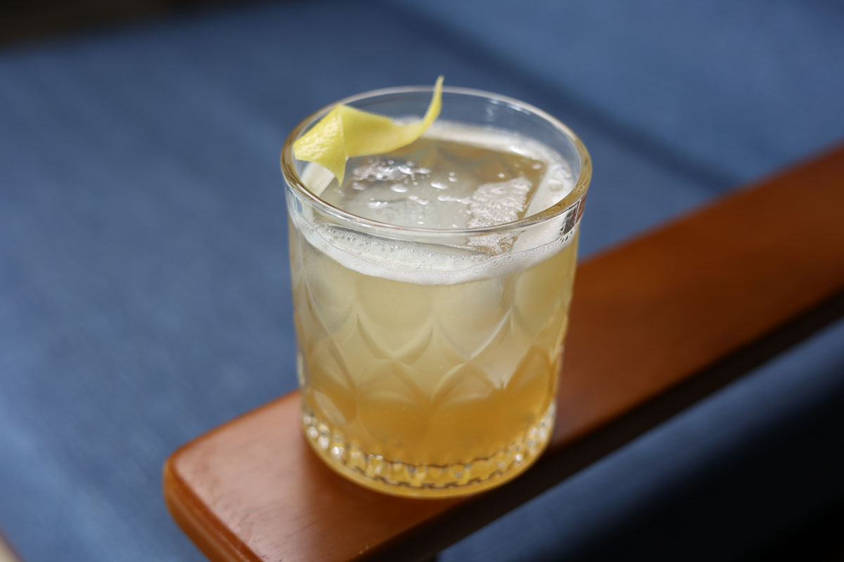 A rocks glass sitting on a wooden armchair containing a cocktail called a 'Gold Rush'.