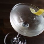 A martini cocktail with a lemon twist rests in a nick and nora glass atop a dark-stained surface.