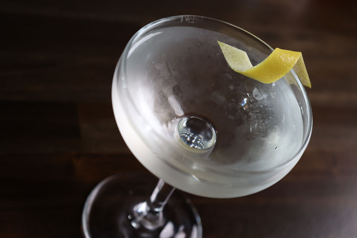 A martini cocktail with a lemon twist rests in a nick and nora glass atop a dark-stained surface.