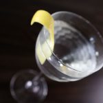 A martini cocktail with a lemon twist rests in a nick and nora glass atop a dark-stained surface.
