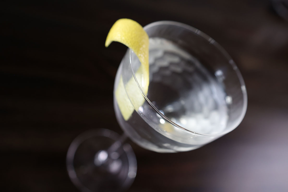 A martini cocktail with a lemon twist rests in a nick and nora glass atop a dark-stained surface.