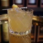 A Penicillin cocktail sits on a gold-colored platter in front of shelves of bourbon bottles.