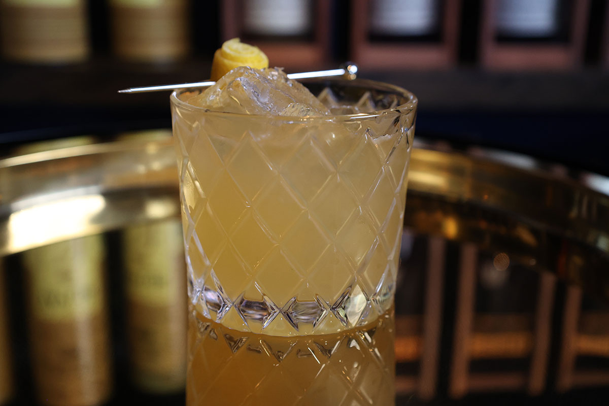 A Penicillin cocktail sits on a gold-colored platter in front of shelves of bourbon bottles.