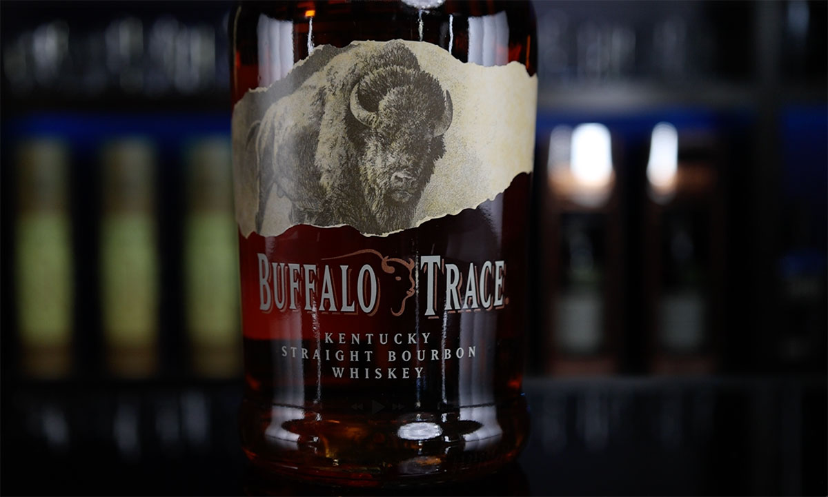 A bottle of Buffalo Trace Bourbon sits on a table in front of shelves of bourbon bottles.