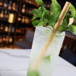 A Mojito cocktail garnished with a mint bunch sits on a white countertop in front of some wood shelves holding distilled spirits bottles.