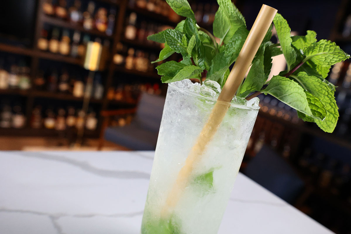 A Mojito cocktail garnished with a mint bunch sits on a white countertop in front of some wood shelves holding distilled spirits bottles.
