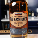A bottle of Costco's Kirkland Signature Old Fashioned Cocktail sits upon a white surface bar top with a back bar in the background.